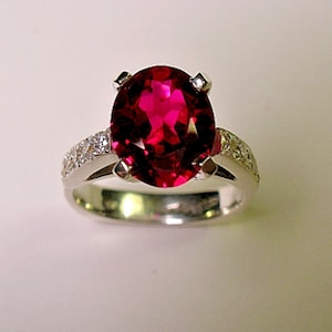 Puede incluir: Un anillo de plata con una piedra preciosa roja grande de forma ovalada rodeada de piedras preciosas blancas más pequeñas.