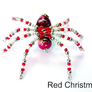 May include: A beaded spider ornament with a red and white color scheme. The spider's body is a marbled red, and its legs are strung with red, white, and silver beads. The text "Red Christmas" is at the bottom.