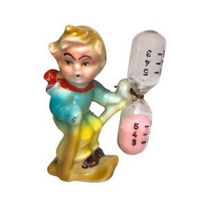 May include: A porcelain figurine of a boy wearing a blue and red outfit, holding a yellow stick and a pink hourglass with the numbers 1, 2, 3, 4, and 5 printed on it.