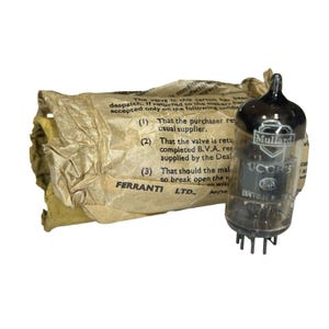 Puede incluir: Una válvula de vacío Mullard UCC85 vintage con una bombilla de vidrio oscuro, junto a un paquete de papel marrón. El tubo tiene el texto "Mullard" y "UCC85" impreso. El paquete de papel tiene texto de Ferranti LTD.
