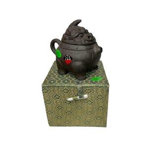 Può includere: Una teiera in ceramica nera a forma di creatura mitologica con un coperchio e un beccuccio. La teiera è appoggiata su una scatola verde e dorata con un motivo decorativo.