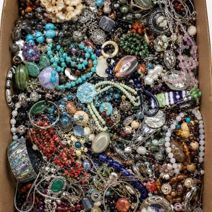 May include: A large cardboard box filled with a diverse collection of costume jewellery, including various beaded necklaces, metal chains, bracelets, rings, and pendants in assorted colours, textures, and materials like glass, stone, and metal.