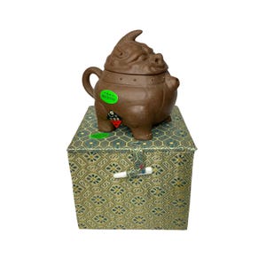Puede incluir: Una tetera de cerámica marrón con forma de criatura mítica con tapa y cuatro patas. La tetera está sentada sobre una caja con estampado verde y dorado con un cierre.