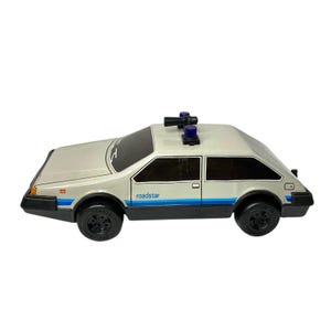 May include: A vintage toy car in a light gray color with black wheels and trim. The car has a blue stripe along the side and the word "roadstar" printed on the side. It features blue and black lights on the roof.