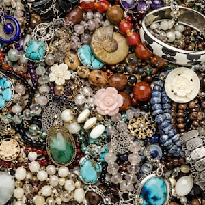 Op de afbeelding: Een dichte verzameling diverse sieraden, waaronder turkoois hangers, kralenkettingen, gesneden bloembedels, een ammonietfossiel, een zwart-witte armband en diverse strengen edelstenen in tinten blauw, groen, bruin en roze.