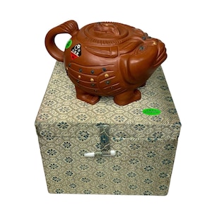 Op de afbeelding: Een bruine theepot van keramiek in de vorm van een mythisch wezen met gouden accenten. De theepot staat op een groen-gouden doos met een sluiting. De doos heeft een label met de tekst "Made in China".