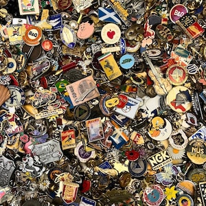 May include: A large collection of assorted metal enamel pins featuring various shapes, logos, and graphic designs including a Ferrari logo, a B&Q sign, a violin, a cartoon bird, and text such as NGK Spark Plugs and Ann Summers Manager.