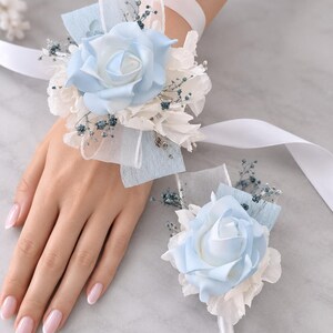 May include: A wrist corsage and a matching boutonniere featuring light blue and white artificial roses, accented with white hydrangea petals, small sprigs of dried blue baby's breath, and light blue ribbon ties on a white satin wrist band.