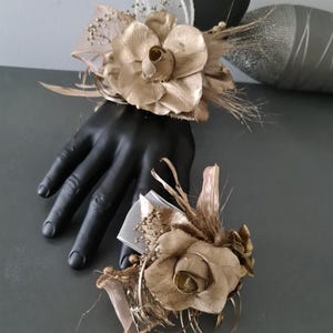 May include: Two gold-colored wrist corsages with floral and feather accents. Each corsage features a large, stylized flower, ribbons, and delicate sprigs. The corsages are displayed on a black mannequin hand, showcasing the design and details.