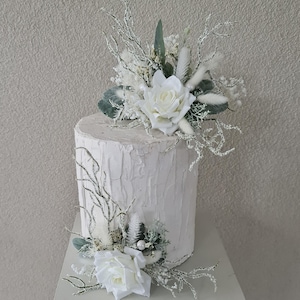 May include: White cake decorated with white and green floral arrangements. The cake is frosted with a white buttercream and has a rustic, wood-like texture. The flowers are made of silk and are arranged in a cluster on top of the cake.
