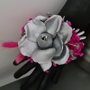 May include: A silver and gray floral corsage with bright pink accents. The corsage features a large, layered flower with a central bud, surrounded by smaller pink flowers and a pink feather. Silver glitter accents add sparkle.