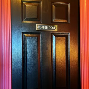 Puede incluir: Una puerta de madera negra con una placa dorada que dice "Powder Room". La puerta tiene un pomo de lat&oacute;n y est&aacute; enmarcada por un marco rojo.