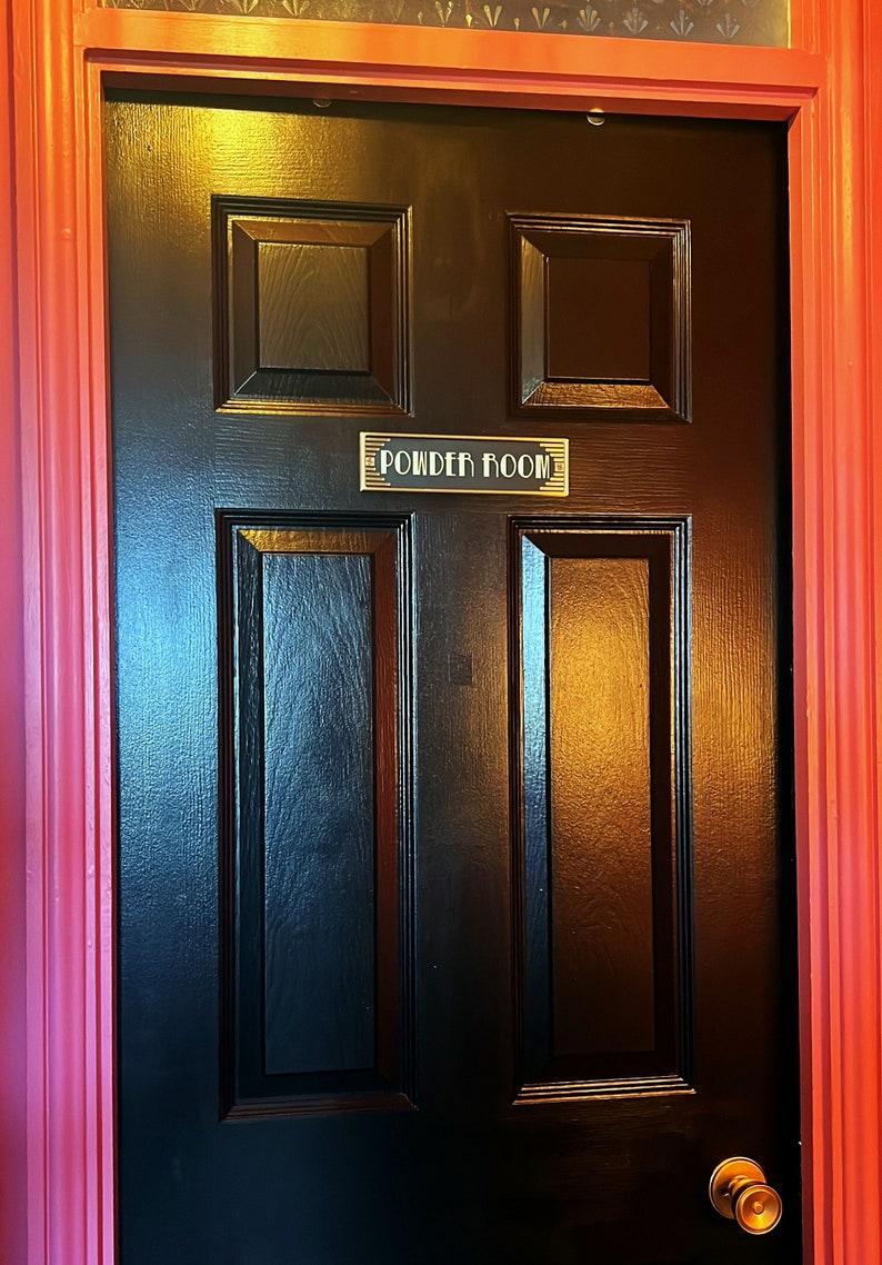 Puede incluir: Una puerta de madera negra con una placa dorada que dice "Powder Room". La puerta tiene un pomo de lat&oacute;n y est&aacute; enmarcada por un marco rojo.