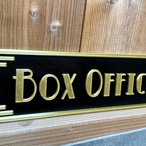 Puede incluir: Un letrero negro con ribete dorado y las palabras "Box Office" en letras doradas.