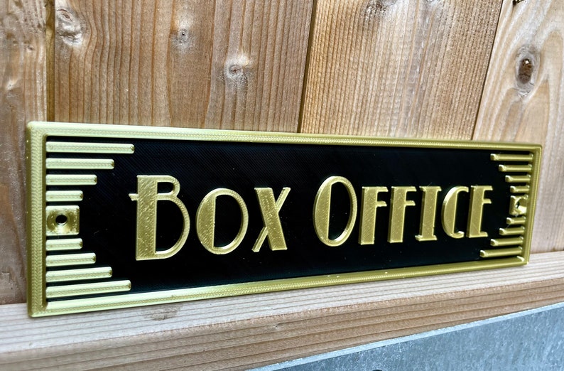 Puede incluir: Un letrero negro con ribete dorado y las palabras "Box Office" en letras doradas.
