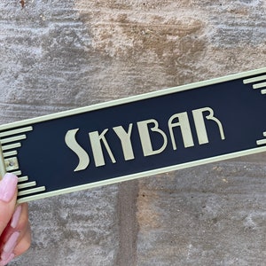 Puede incluir: Un letrero de metal negro y dorado con la palabra "Skybar" en letras doradas.