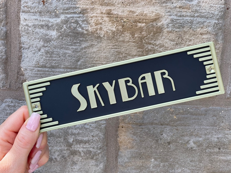 Puede incluir: Un letrero de metal negro y dorado con la palabra "Skybar" en letras doradas.