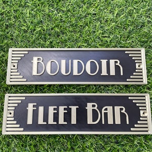 Puede incluir: Dos letreros rectangulares negros con bordes dorados y letras blancas. El letrero superior dice "BOUDOIR" y el letrero inferior dice "FLEET BAR".