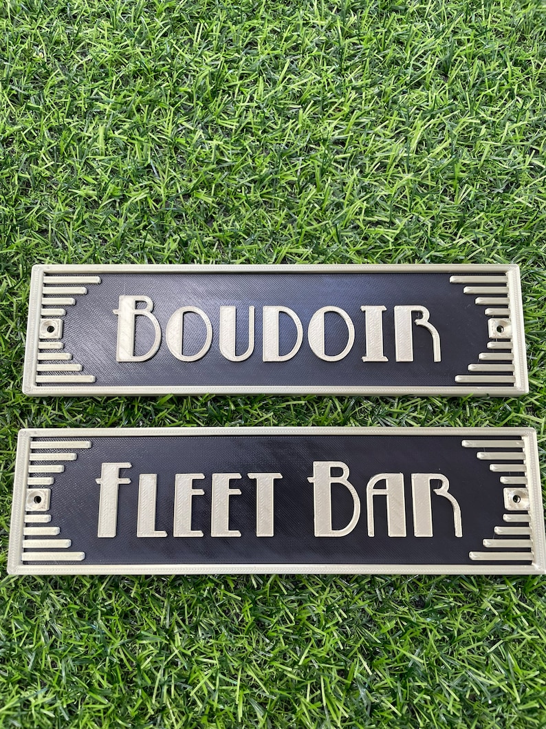 Puede incluir: Dos letreros rectangulares negros con bordes dorados y letras blancas. El letrero superior dice "BOUDOIR" y el letrero inferior dice "FLEET BAR".