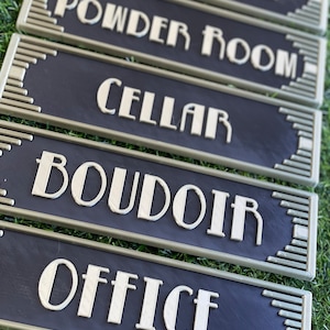 Puede incluir: Cinco letreros rectangulares negros y dorados con letras blancas. Los letreros dicen "Elvis Suite", "Powder Room", "Cellar", "Boudoir" y "Office".