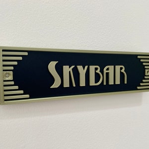 Puede incluir: Un letrero negro con borde dorado y la palabra "SKYBAR" en letras doradas.