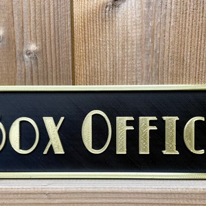 Puede incluir: Un letrero negro con ribete dorado y las palabras "Box Office" en letras doradas.