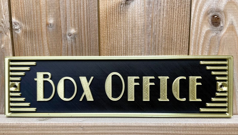 Puede incluir: Un letrero negro con ribete dorado y las palabras "Box Office" en letras doradas.