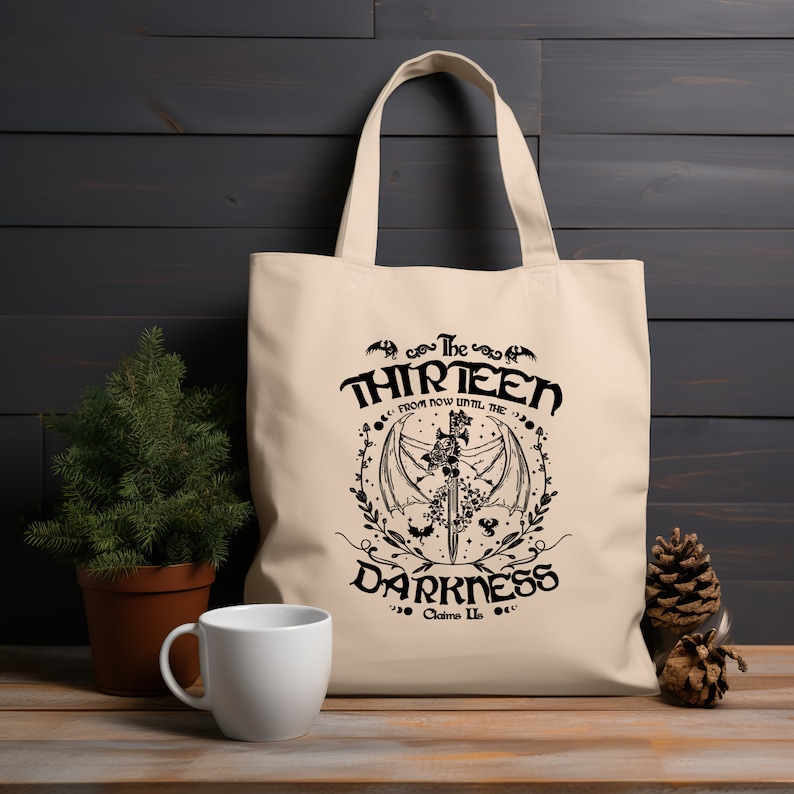 May include: A beige tote bag with black text and a graphic design. The text reads "The Thirteen From Now Until The Darkness Claims Us".