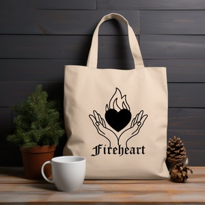 May include: A beige tote bag with a black graphic of two hands holding a heart with flames. The text "Fireheart" is printed below the graphic.
