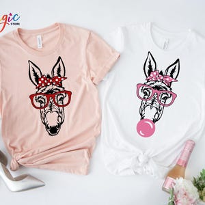 May include: Two t-shirts with a graphic of a donkey wearing glasses and a red and white polka dot headband. One shirt is pink and the other is white. The donkey on the white shirt is blowing a pink bubble.