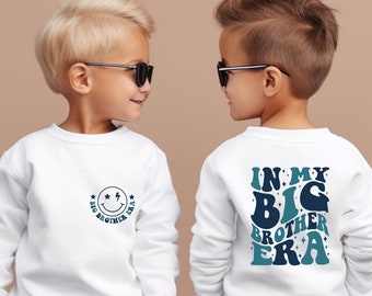 May include: Two children wearing white sweatshirts with blue and green text. The front of the sweatshirt says "Big Brother Era" with a smiley face. The back of the sweatshirt says "In My Big Brother Era".