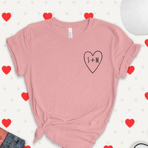 May include: A pink t-shirt with a black heart graphic that says "S+M" on it. The shirt is on a white background with red hearts.