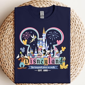 Puede incluir: Camiseta azul marino con un dise&ntilde;o colorido de Disneyland. El dise&ntilde;o incluye el logotipo de Disneyland, Mickey Mouse y el texto "The happiest place on earth - EST. 1955". La camiseta est&aacute; doblada sobre una superficie tejida.