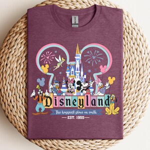 Puede incluir: Una camiseta granate con un gr&aacute;fico colorido de Disneyland. El dise&ntilde;o incluye el logotipo de Disneyland, un castillo, fuegos artificiales y varios personajes de Disney. El texto dice "Disneyland" y "The happiest place on earth - EST. 1955."