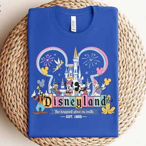Puede incluir: Camiseta azul real con un gr&aacute;fico colorido de Disneyland. El dise&ntilde;o incluye el logotipo de Disneyland, un castillo, fuegos artificiales y varios personajes de Disney. El texto dice "Disneyland, The happiest place on earth, EST. 1955."