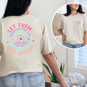 May include: A beige t-shirt with a colorful graphic on the back that says "Let them misunderstand you, judge you, gossip about you, never doubt your worth, you are beautiful, keep shining!" The graphic features a daisy flower and other floral elements.