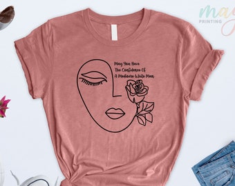 May include: A pink t-shirt with a black line drawing of a face with closed eyes and a rose. The text "May You Have The Confidence Of A Mediocre White Man" is printed on the shirt.