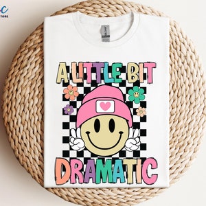 May include: White t-shirt with a colorful graphic of a smiley face wearing a pink beanie and the text "A little bit dramatic".