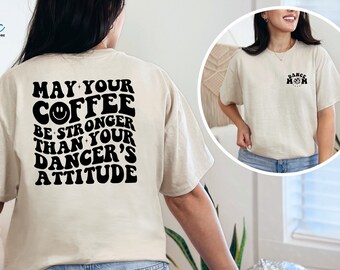 Op de afbeelding: Een beige T-shirt met zwarte tekst die "May your coffee be stronger than your dancer's attitude" luidt. De tekst is in een retro-lettertype met een smiley in het woord "coffee". De achterkant van het shirt is te zien. Een tweede beige T-shirt is te zien met zwarte tekst die "Dance Mom" luidt met een smiley in het woord "Mom".