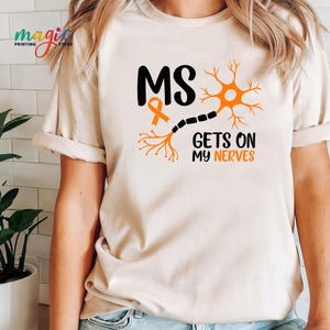 May include: Beige t-shirt with the text "MS GETS ON MY NERVES" in black and orange. The design includes an orange ribbon and a stylized neuron graphic. The shirt has rolled-up sleeves.