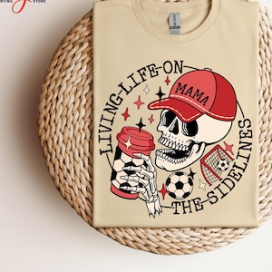 May include: A beige t-shirt featuring a graphic of a skull wearing a red baseball cap with the word "Mama" on it. The skull is holding a coffee cup and a football. The text "Living Life On The Sidelines" is printed around the skull.