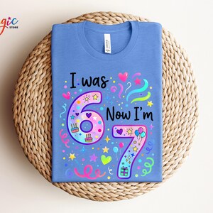May include: A blue t-shirt featuring the text "I was 6 Now I'm 7" in a vibrant design. The design incorporates the numbers 6 and 7, hearts, stars, balloons, and confetti. The shirt is presented on a woven, circular surface.