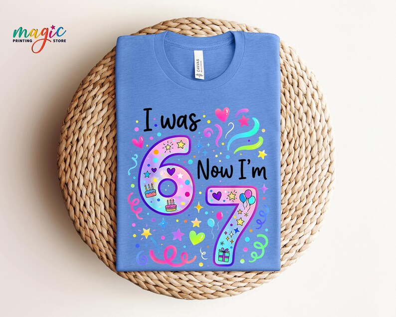 May include: A blue t-shirt featuring the text "I was 6 Now I'm 7" in a vibrant design. The design incorporates the numbers 6 and 7, hearts, stars, balloons, and confetti. The shirt is presented on a woven, circular surface.