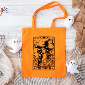 May include: An orange tote bag featuring a black graphic print of a person wearing a wide-brimmed hat, holding a black cat, surrounded by floral elements and moon phases, with the text THE WITCH printed at the bottom of the rectangular frame.