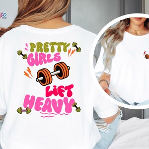 Puede incluir: Camiseta blanca con el texto "PRETTY GIRLS LIFT HEAVY" en rosa y verde, junto con un gr&aacute;fico de barra. La camiseta tambi&eacute;n presenta un peque&ntilde;o gr&aacute;fico de barra en la parte delantera. El dise&ntilde;o incluye acentos naranjas y amarillos.