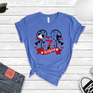 以下が含まれることがあります： ミッキーマウスの頭のデザインが施された青いTシャツ。頭は2つの円形で、中に数字の7が入っています。「my 7th birthday」という文字が赤いリボンに書かれています。
