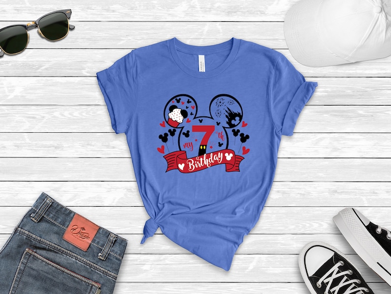 以下が含まれることがあります： ミッキーマウスの頭のデザインが施された青いTシャツ。頭は2つの円形で、中に数字の7が入っています。「my 7th birthday」という文字が赤いリボンに書かれています。
