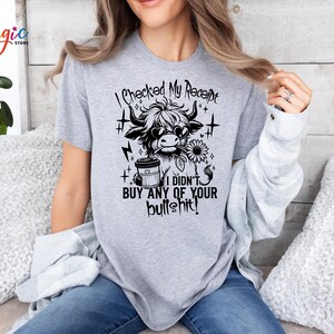 May include: A light gray t-shirt featuring a black and white graphic of a Highland cow wearing sunglasses and holding a sunflower. The text "I checked my receipt, I didn't buy any of your bullshit!" is printed above and below the graphic.