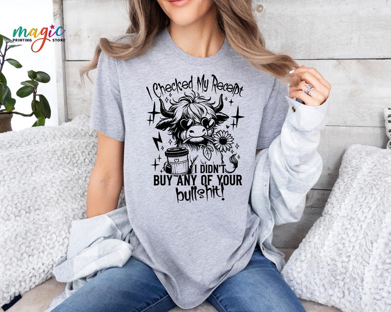 May include: A light gray t-shirt featuring a black and white graphic of a Highland cow wearing sunglasses and holding a sunflower. The text "I checked my receipt, I didn't buy any of your bullshit!" is printed above and below the graphic.