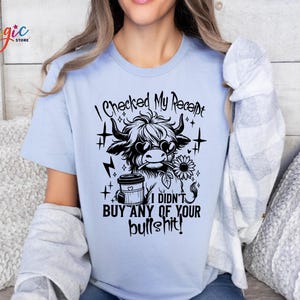 May include: Light blue t-shirt with a black and white graphic of a cow wearing sunglasses and holding a flower. Text on the shirt reads "I CHECKED MY RECEIPT I DIDN'T BUY ANY OF YOUR BULLSHIT!" The shirt has short sleeves.